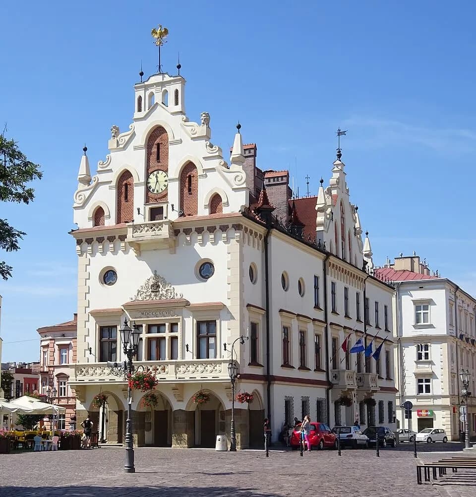 A historic white building with ornate architectural details, a clock tower, and a golden eagle finial stands prominently in a town square under a clear blue sky, with other buildings and a few cars visible in the background.